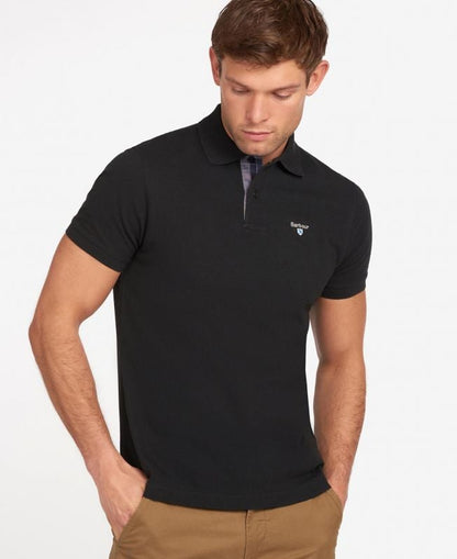 A man wearing a black Barbour Tartan Pique polo shirt with a two-button placket and a collar. The shirt features a ribbed cuff and is detailed with tartan pattern to the inner neckline and a Barbour crest logo on the chest.