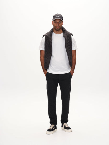 A person wearing a white t-shirt, dark grey knitted waistcoat, black trousers, and black shoes, standing against a white background.